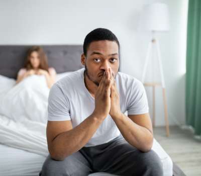 young-black-guy-feeling-unhappy-sitting-on-bed-deep-in-thought-his-disappointed-wife-on-background